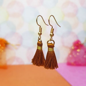 Könnte beinhalten: Ein Paar goldene Quastenohrringe mit braunen Quasten. Die Ohrringe befinden sich auf einem rosa und orangefarbenen Hintergrund.