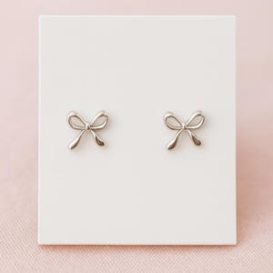 Peut inclure: Une paire de boucles d'oreilles clous en forme de nœud papillon argenté. Les boucles d'oreilles sont présentées sur un fond blanc. Les nœuds ont un design simple et élégant, avec des lignes courbes et une finition polie.