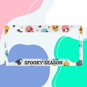 Puede incluir: Marco de matrícula blanco con temática de temporada de miedo. El marco está decorado con calaveras y estrellas de colores. El texto "SPOOKY SEASON" está impreso en letras negras en el marco.