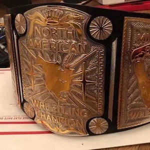 Könnte beinhalten: Ein gold-silberner Wrestling-Meisterschaftsgürtel mit den Worten "North American Wrestling Champion" und "Mid South Wrestling". Der Gürtel hat einen schwarzen Lederriemen und aufwendige Details.