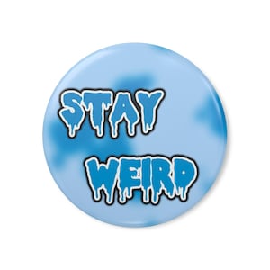 Puede incluir: Un botón redondo azul claro con las palabras "STAY WEIRD" en letras azules goteantes con contorno blanco. El fondo tiene un efecto nublado, similar a una acuarela.