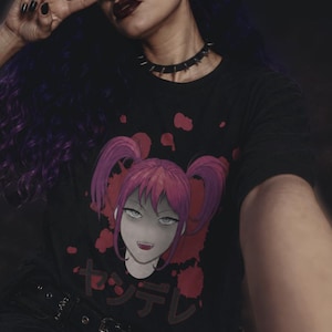 Może przedstawiać: Czarny t-shirt z różowowłosą postacią z anime i japońskim tekstem "Yandere". Grafika otoczona jest czerwonymi plamami. Widoczny jest czarny choker z ćwiekami.