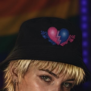 May include: Black bucket hat featuring a pink and blue heart design split by a lightning bolt. The heart is surrounded by floral accents in pink and blue. The hat is made of a soft material.