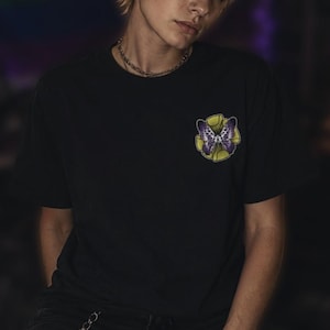 May include: Black t-shirt featuring a butterfly design. The butterfly is purple and white, set against a yellow and green floral background. The shirt is a classic crew neck style.