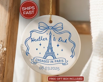 May include: A white ceramic ornament with a blue Eiffel Tower design, the names "Heather & Erik", and the date 08.03.2025. The ornament is round and has a gold string for hanging. The words "Engaged in Paris" are also included.