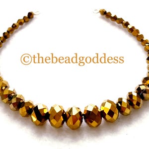 May include: A curved strand of faceted metallic gold beads arranged in a graduated size pattern, with the largest beads in the center and smaller beads toward the ends. The text "©thebeadgoddess" is printed in gold in the center of the arc.