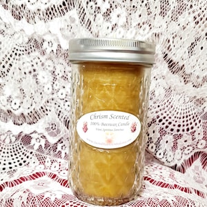 May include: A clear glass jar candle with a gold lid and a label that reads "Chrism Scented 100% Beeswax Candle Veni Spiritus Sanctus www.sacredheartsunited.etsy.com".
