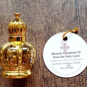 May include: A gold-coloured crown-shaped glass bottle with a matching cap, accompanied by a round white tag featuring a cross symbol and the text Blessed Anointing Oil from the Holy Land, Frankincense, Myrrh, & Spikenard, Net wt. 15ml.