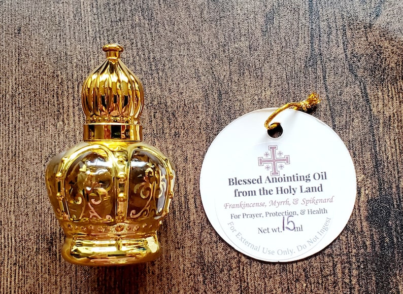 May include: A gold-coloured crown-shaped glass bottle with a matching cap, accompanied by a round white tag featuring a cross symbol and the text Blessed Anointing Oil from the Holy Land, Frankincense, Myrrh, & Spikenard, Net wt. 15ml.
