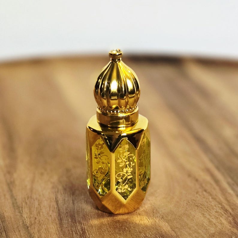 May include: A small, decorative glass perfume bottle with a yellow-tinted body featuring an etched floral pattern. The bottle is topped with a shiny, metallic gold cap shaped like a rounded dome with vertical ridges and a small finial at the top.