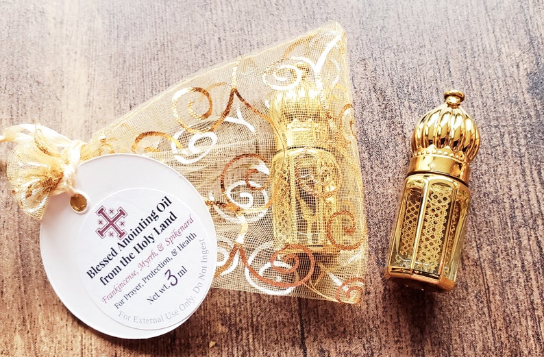 May include: A small glass bottle with a gold-coloured ornate cap and metal lattice overlay, accompanied by a gold organza drawstring pouch and a round white tag labelled Blessed Anointing Oil from the Holy Land, Net wt. 3 ml.