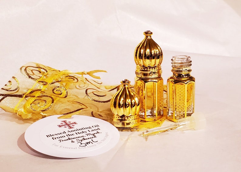 May include: Two small glass bottles with gold-coloured ornate caps, one with the cap removed, alongside a clear glass dropper. A round white tag reads Blessed Anointing Oil from the Holy Land, Frankincense, Myrrh, & Spikenard, 3ml. A gold organza bag.