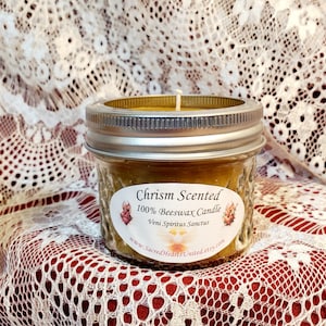 May include: A small glass jar candle with a gold lid. The candle is made of 100% beeswax and is scented with chrism. The label reads "Chrism Scented 100% Beeswax Candle Veni Spiritus Sanctus www.SacredHeartsUnited.etsy.com".