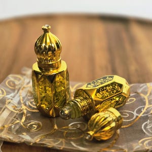 May include: Two small, gold-toned perfume bottles with faceted glass bodies and ornate, dome-shaped caps. One bottle stands upright while the other lies on its side with its cap removed, revealing a rollerball applicator and intricate floral patterns.