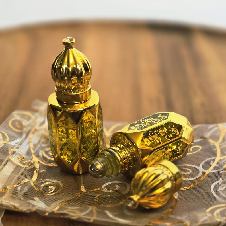 May include: Two small, gold-toned perfume bottles with faceted glass bodies and ornate, dome-shaped caps. One bottle stands upright while the other lies on its side with its cap removed, revealing a rollerball applicator and intricate floral patterns.