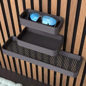 May include: Three-tiered gray storage shelves with a textured surface, mounted on a wooden wall. The top shelf holds blue-tinted sunglasses. The shelves are designed for organizing small items.