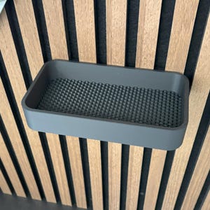 May include: A gray rectangular shelf with a textured interior, mounted on a wooden slatted wall. The shelf is a solid, muted gray color, and the interior has a grid-like pattern.