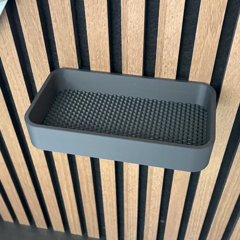 May include: A gray rectangular shelf with a textured interior, mounted on a wooden slatted wall. The shelf is a solid, muted gray color, and the interior has a grid-like pattern.