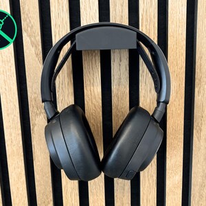 Könnte beinhalten: Schwarzer Over-Ear-Kopfhörer, der an einer schwarzen Wandhalterung hängt. Die Kopfhörer hängen an einer Holzwand mit vertikalen hellbraunen und schwarzen Streifen. Ein grüner Kreis mit einem durchgestrichenen Bohrer-Symbol befindet sich in der oberen linken Ecke.