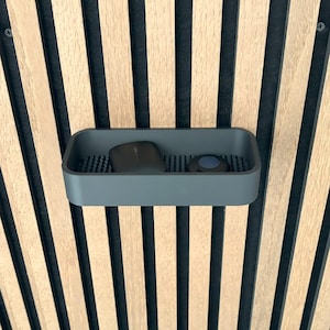 May include: A gray rectangular tray with rounded corners, holding a dark-colored object and a small round object. The tray sits against a backdrop of vertical wooden slats alternating with black stripes.