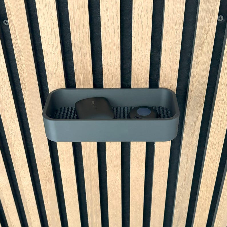 May include: A gray rectangular tray with rounded corners, holding a dark-colored object and a small round object. The tray sits against a backdrop of vertical wooden slats alternating with black stripes.