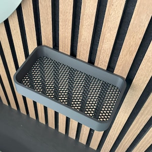 May include: A rectangular, gray soap dish with a perforated black insert. The dish is mounted on a wall with alternating vertical wooden and black panels. The design is modern and minimalist.