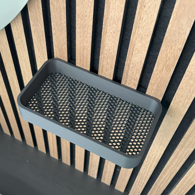 May include: A rectangular, gray soap dish with a perforated black insert. The dish is mounted on a wall with alternating vertical wooden and black panels. The design is modern and minimalist.