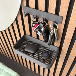 May include: A gray key holder with a shelf and hooks, mounted on a wooden wall. Several sets of keys hang from the hooks, and two small electronic devices rest on the shelf. The wall has vertical wooden slats.
