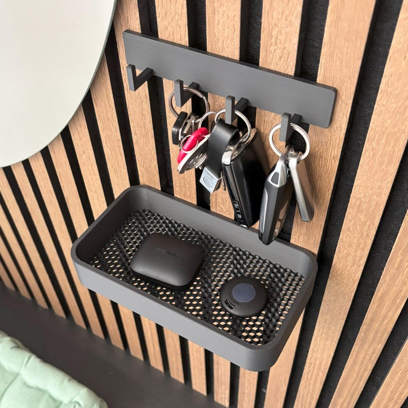 May include: A gray key holder with a shelf and hooks, mounted on a wooden wall. Several sets of keys hang from the hooks, and two small electronic devices rest on the shelf. The wall has vertical wooden slats.