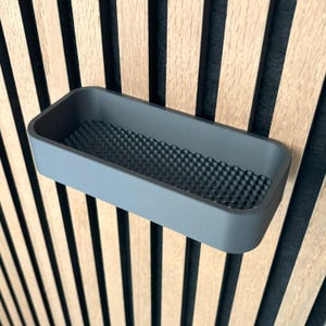 May include: A rectangular, gray storage tray with a textured, geometric pattern on the inside. The tray is mounted on a wooden wall with vertical, alternating light and dark wood panels.