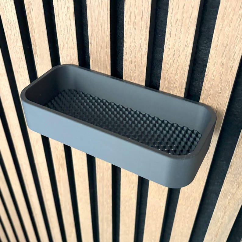 May include: A rectangular, gray storage tray with a textured, geometric pattern on the inside. The tray is mounted on a wooden wall with vertical, alternating light and dark wood panels.