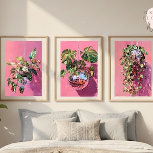 Könnte beinhalten: Drei gerahmte Wandkunstdrucke mit Pflanzen in Discokugel-Pflanzgefäßen vor einem rosa Hintergrund. Die Drucke befinden sich in Holzrahmen und werden über einem Bett mit Kissen und einer Bettdecke ausgestellt. Die Kunst trägt die Bezeichnung "Dreamy Flowers Wall Art."