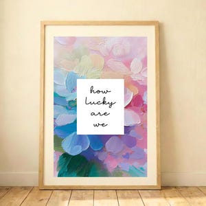 Puede incluir: Una impresión artística enmarcada con un diseño floral abstracto en tonos rosa, azul y morado. La obra de arte tiene un aspecto texturizado y pintado. Un cuadrado blanco en el centro muestra el texto "how lucky are we" en una fuente manuscrita. El marco es de madera clara.