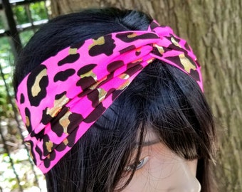 以下が含まれることがあります： 黒と金色のヒョウ柄模様の明るいピンク色のヘアバンド。ヘアバンドは上部で結ばれており、黒髪の人が着用しています。背景はぼやけており、屋外の環境を示唆しています。