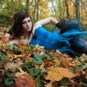 May include: A woman wearing a blue corset and a black and blue tulle skirt lies on a bed of autumn leaves.