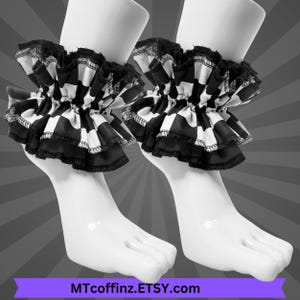 May include: A pair of black and white checkered ruffled ankle cuffs. The cuffs are made of fabric and have a layered design with a black trim. The cuffs are displayed on white mannequin feet. The text "MTcoffinz.ETSY.com" is at the bottom.