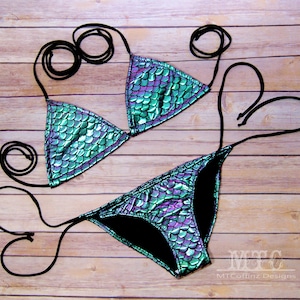 May include: A green and purple iridescent bikini with a mermaid scale pattern. The bikini top is a triangle style with adjustable straps. The bikini bottom is a cheeky style with a high cut leg.