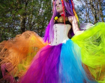 May include: A woman wearing a white corset with colorful tulle skirt. Her hair is styled with colorful dreadlocks. She is standing in a forest setting.