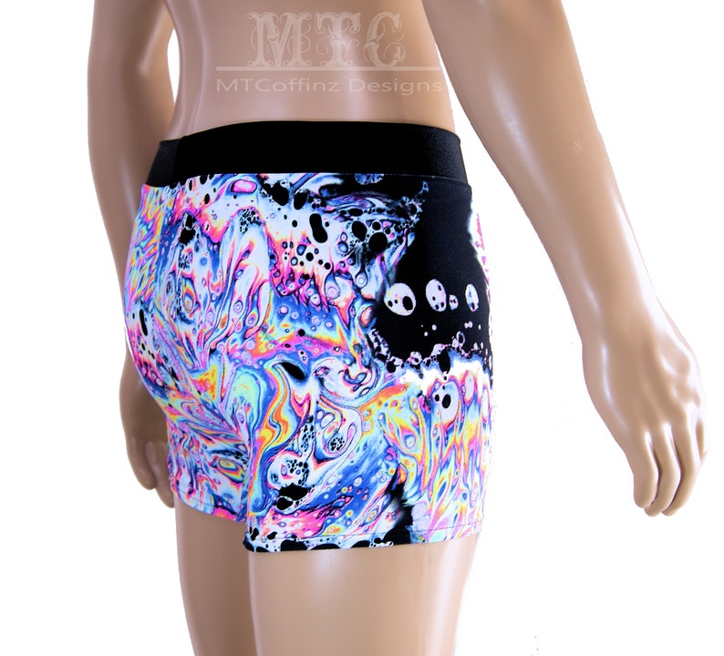 Puede incluir: Pantalones cortos con cintura negra y un vibrante patr&oacute;n arremolinado en tonos de azul, rosa, amarillo y blanco. Los pantalones cortos tienen un panel negro con dise&ntilde;os circulares blancos. El texto "MTCoffinz Designs" es visible.