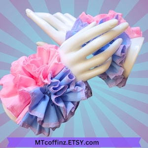 May include: A pair of pink and blue ruffled fabric wrist cuffs. The cuffs are made of two layers of fabric, with the top layer being pink and the bottom layer being blue. The cuffs are tied together with a ribbon.