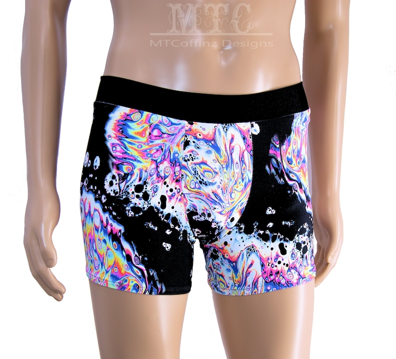 Puede incluir: B&oacute;xer negro con un vibrante dise&ntilde;o en espiral en tonos rosa, azul, amarillo y blanco, que recuerda el efecto de una burbuja de jab&oacute;n. La cintura es de color negro liso. El texto "MTCoffinz Designs" es visible.