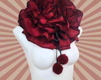 May include: A large, red, ruffled collar with black ribbon ties and two red pom-poms.  The collar is made of fabric and is a statement piece.