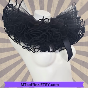 May include: A black ruffled collar made of tulle with a black satin ribbon tied at the back. The collar is on a white mannequin.