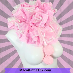 May include: A pink ruffled collar with white trim and two fluffy pink pom-poms attached. The collar is displayed on a white mannequin bust.