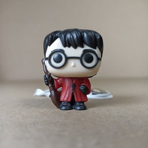 May include: A small plastic figurine of Harry Potter from the Harry Potter series. He is wearing a red robe and holding a brown broomstick. He has black hair and round glasses.