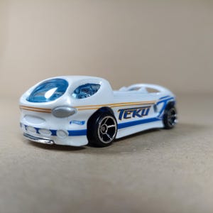 May include: White toy car with blue and gold accents. The car has a clear windshield and the word "TEKU" printed on the side.