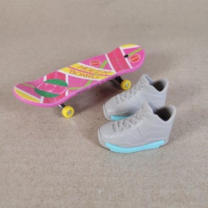 May include: A miniature skateboard with a pink and yellow design, and the words "HOVER BOARD". Beside it are two grey toy trainers with light blue soles. The skateboard has yellow wheels.