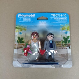 May include: Playmobil 71507 Bride and Groom set with 11 pieces. The bride is wearing a white dress and holding a bouquet of red and pink flowers. The groom is wearing a gray suit and holding a red heart. The set is packaged in a clear plastic blister pack with a blue and white background.