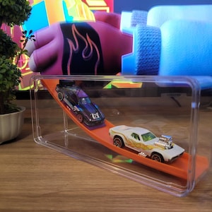 Könnte beinhalten: Eine transparente Acryl-Vitrine mit einer orangefarbenen Rennstrecke und zwei Spielzeugautos. Eines ist schwarz und lila, das andere weiß mit orangefarbenen Flammen. Die Vitrine steht auf einer Holzoberfläche.