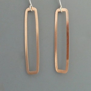 Puede incluir: Un par de pendientes rectangulares de cobre con un gancho de plata. Los pendientes son simples y de diseño moderno.